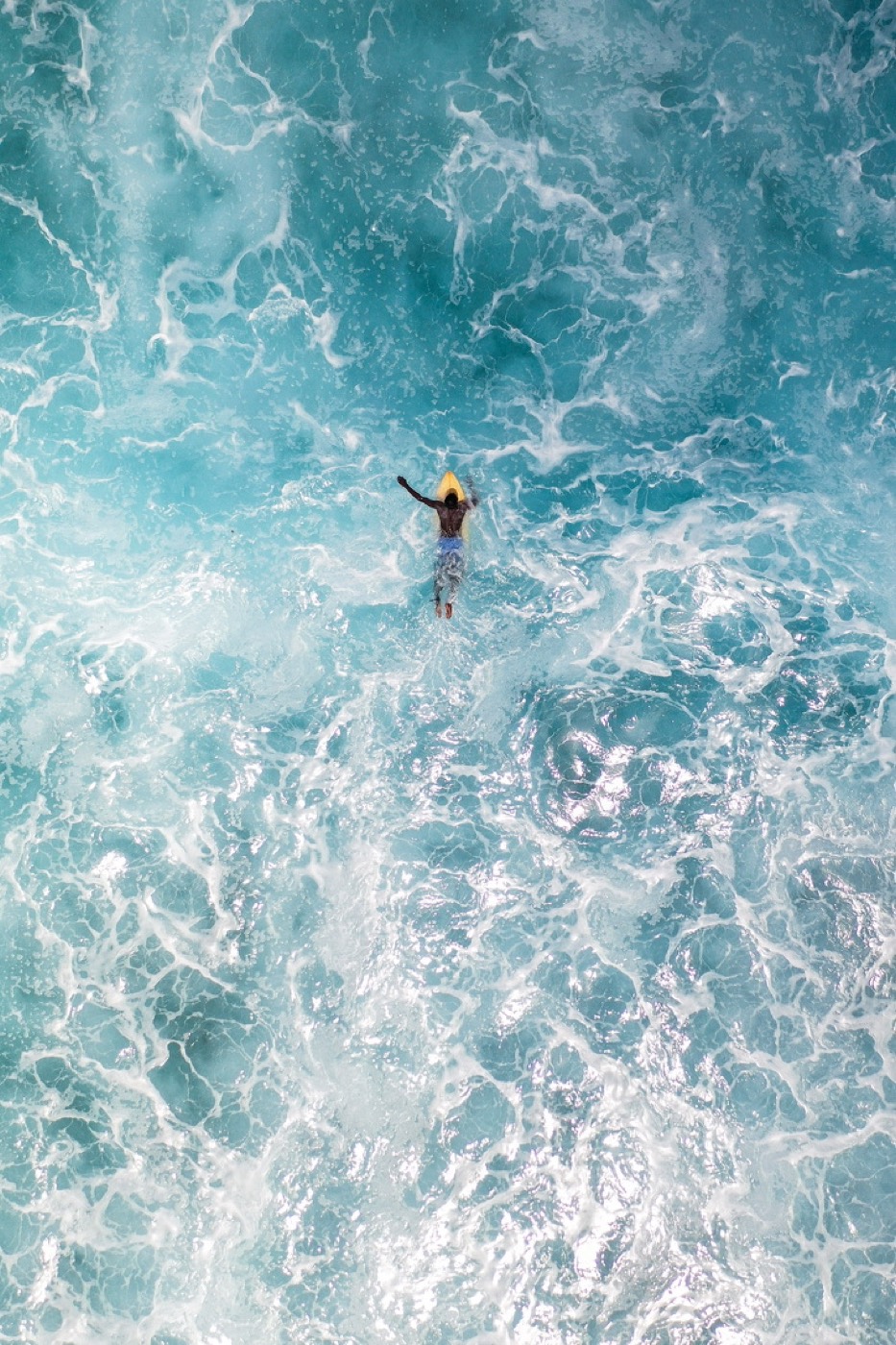 An aerial view of a surfer paddling out through white water over a turquoise reef